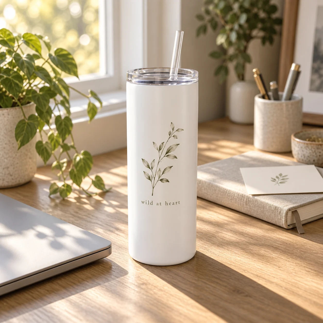A white botanical skinny tumbler on an oak window desk beside a closed laptop and a note